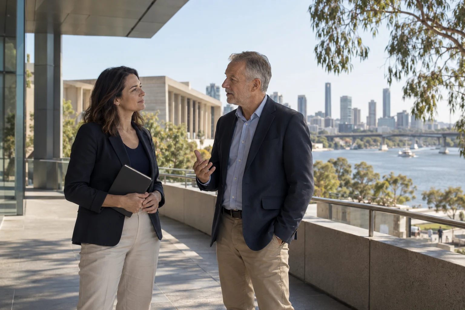 Two colleagues in conversation on a shaded terrace overlooking an Australian river city.